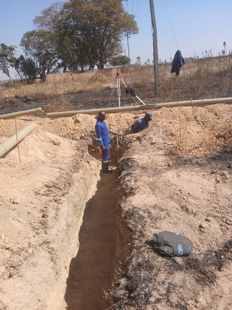 Surveying level on a tripod beside an open sewer trench during sewer setting out, with pipes laid out along the trench