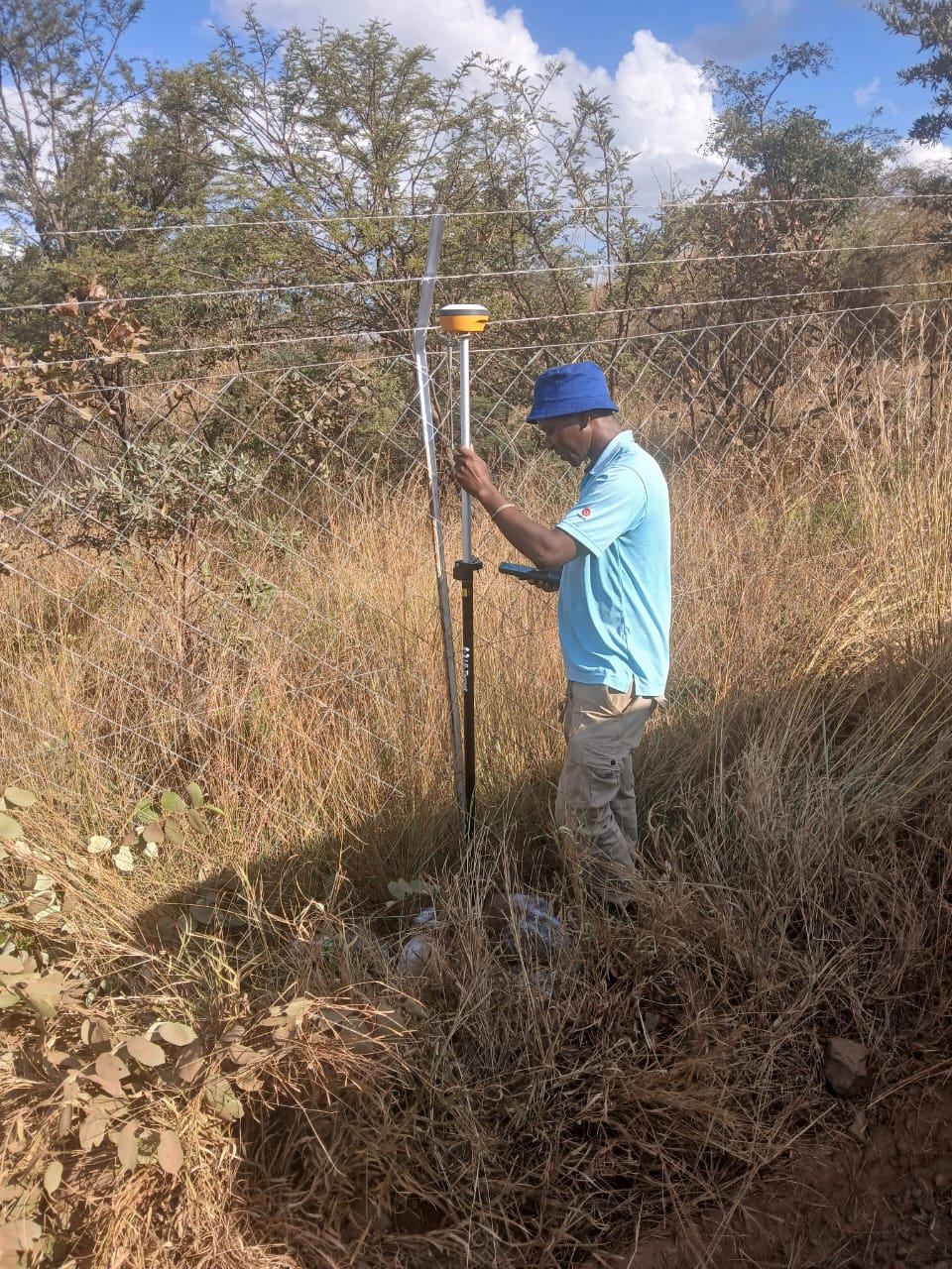 Surveyor with a GNSS pole and handheld controller beside a boundary fence during a topographical survey