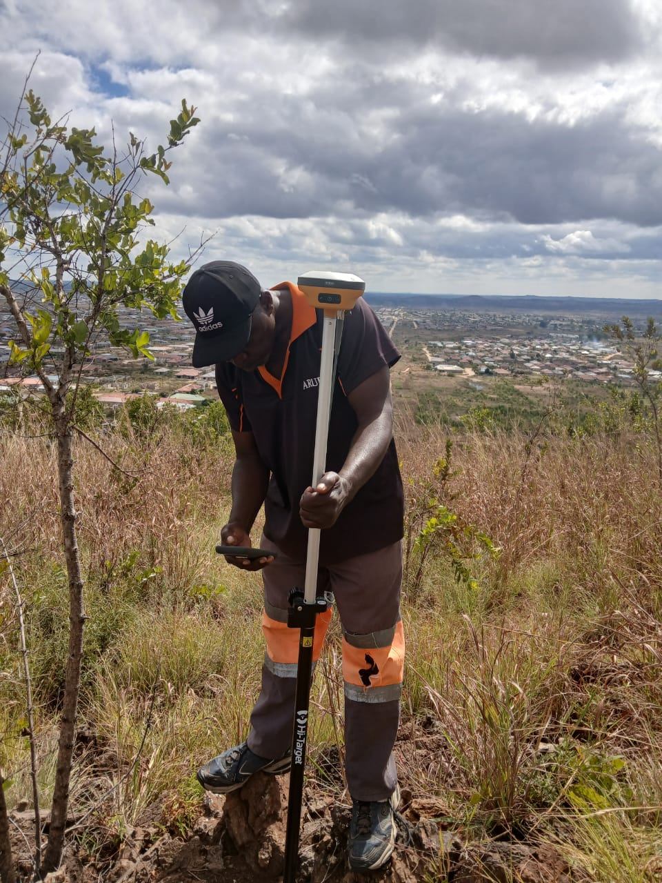 Surveyor using a GNSS pole for a topographical and boundary survey on a hillside overlooking a residential area